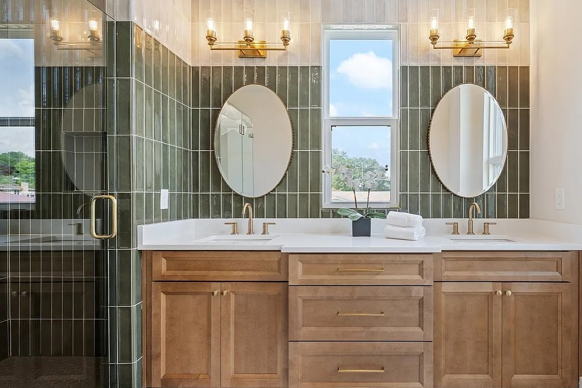 Primary bathroom lined in green glazed tile, with twin oval mirrors and brass sconces above an oak vanity.