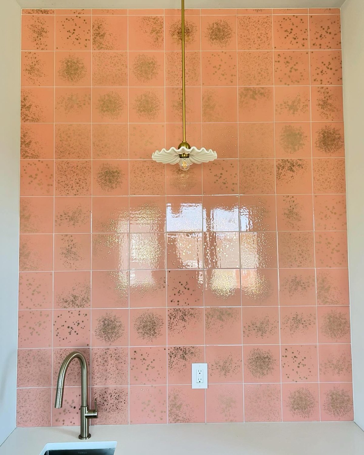 A bathroom wall tiled floor to ceiling in speckled pink tile, with a brass scalloped shower rose.